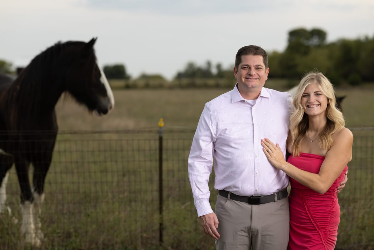 Lauren and Kyle smiling together beside a pasture fence, a horse looking on.